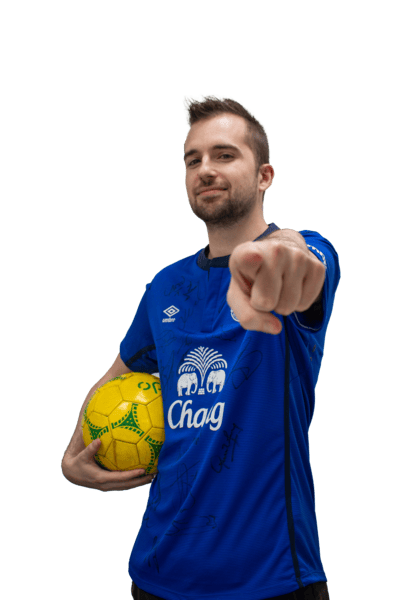 A man wearing the blue Everton Football Club home shirt. He is holding a yellow ball under his arm and pointing at the camera. 