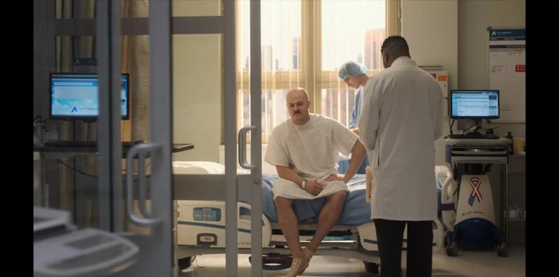 A patient sitting on a hospital bed in a hospital gown, with a doctor standing next to him discussing his treatment.
