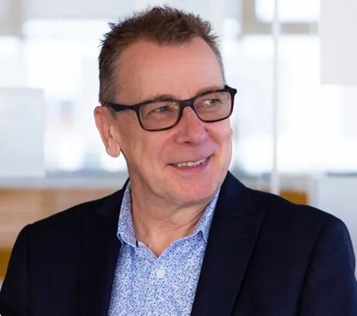 A white middle aged male in glasses. He is wearing a blue patterned shirt and navy suit jacket