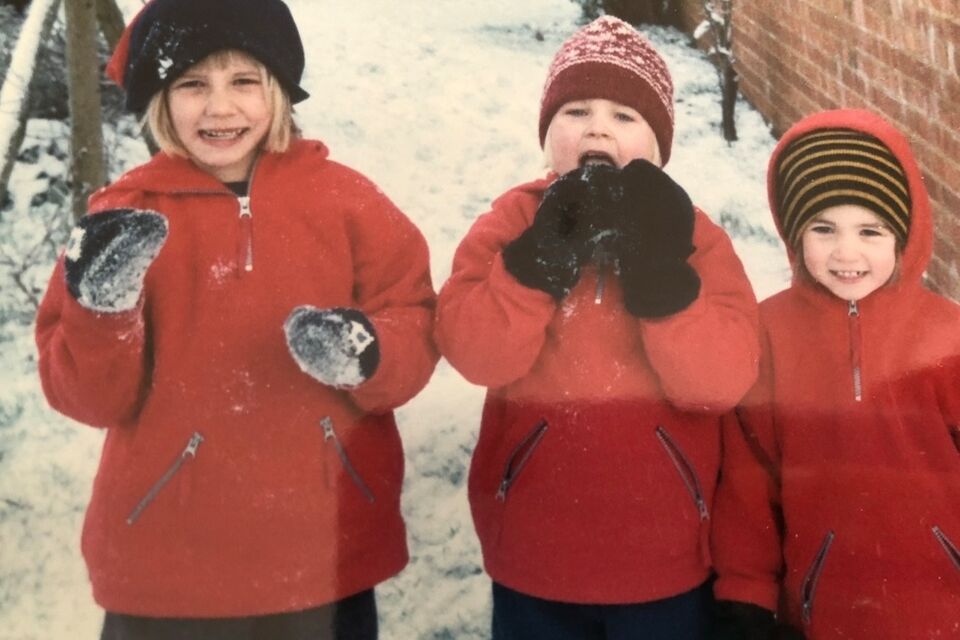 three children wearing red coats and wooly hats with snow behind them