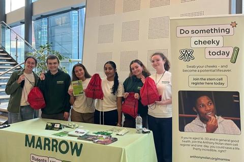 A group of six young people are stood behind a table holding up leaflets and bags. They are running a donor recruitment event.