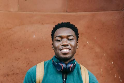 A black male smiling. He is wearing a green jumper and has a light brown backpack on his shoulders. He has black headphones around his neck, he is clean shaven and has low cut black afro hair.