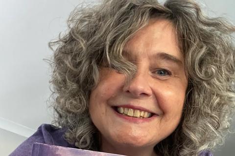 Michelle smiling and holding a copy of her book, she has grey curly hair and a purple t-shirt