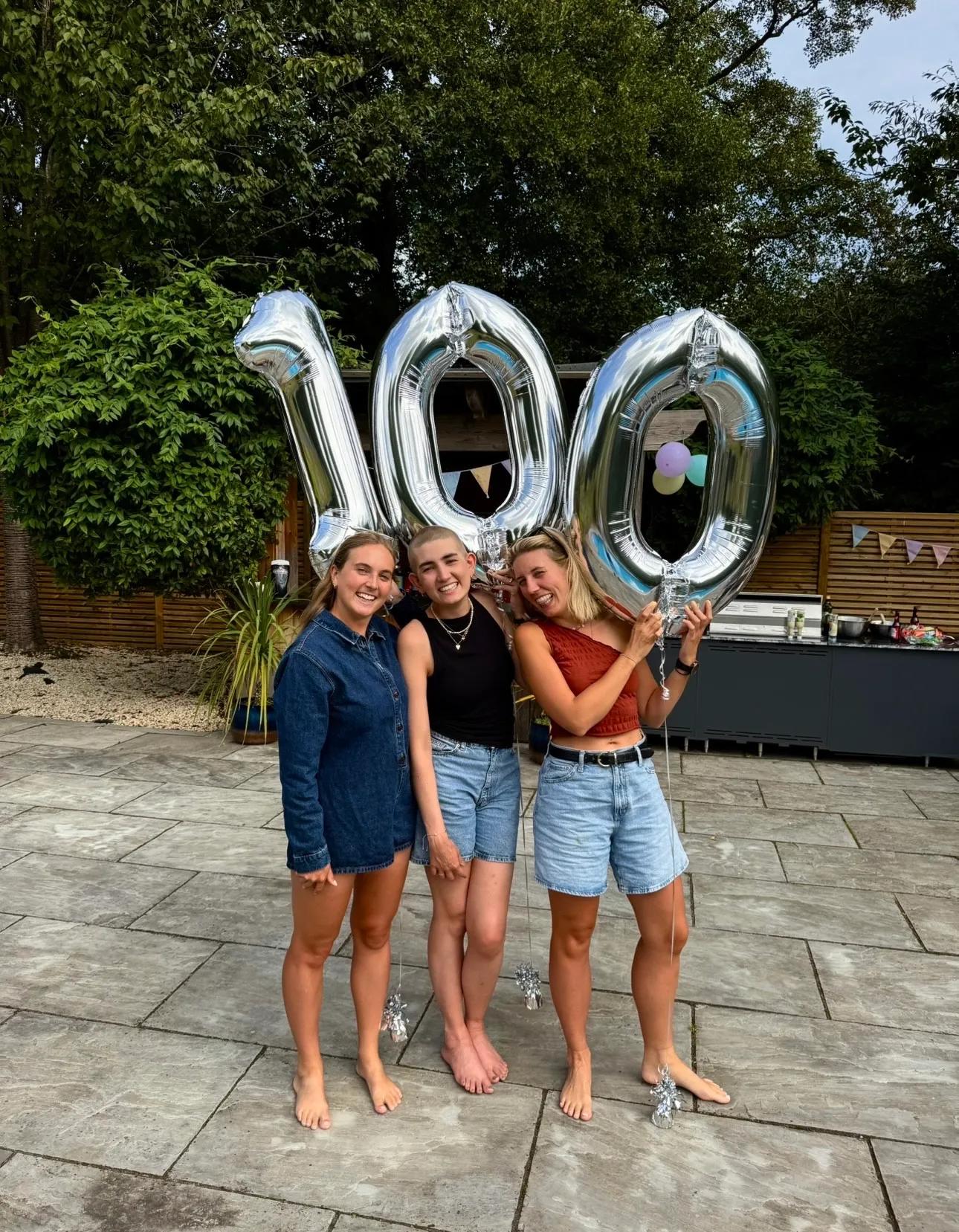three young women standing together under silver balloons that read '100'