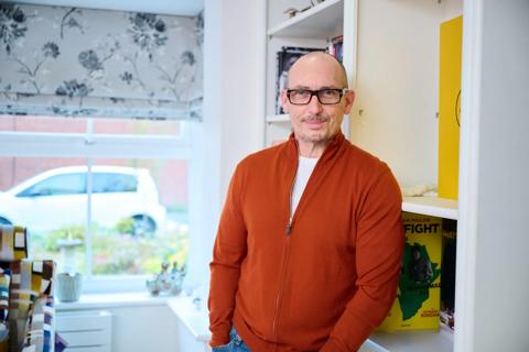 A middle aged white man smiling wearing black thick rim glasses and an orange jumper leaning against some display shelves at home