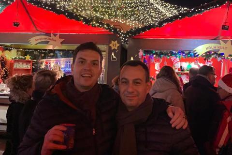 two men stand with arms around each other holding a beer with  a christmas market behind them