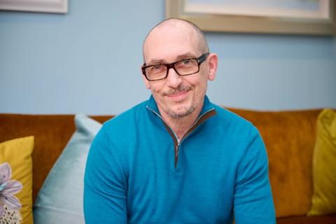 A middle aged white man smiling wearing black thick rim glasses and a blue jumper sat on his sofa at home
