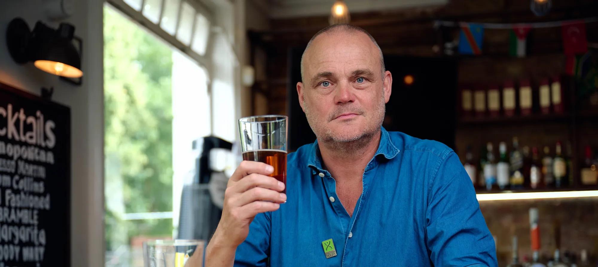 Comedian Al Murray holding half a pint of beer 