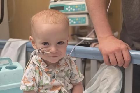 A baby boy sits in a hospital cot playing with some toys. He has  dinosaur pyjamas on and a feeding tube in his nose.