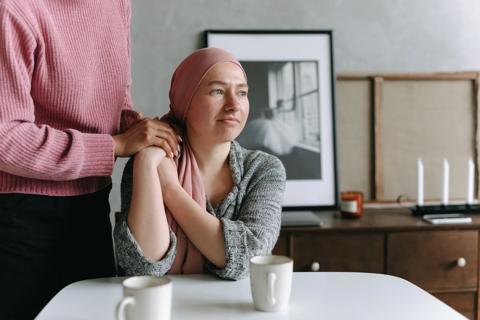 A white woman wearing a headscarf sitting at a table looking into the distance whilst holding hands with another woman to the side 