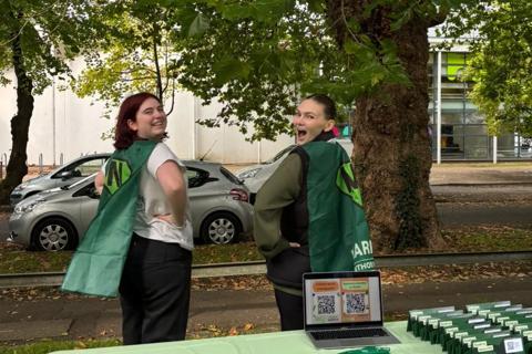 two young women posing in capes behind a Marrow donor recruitment stall