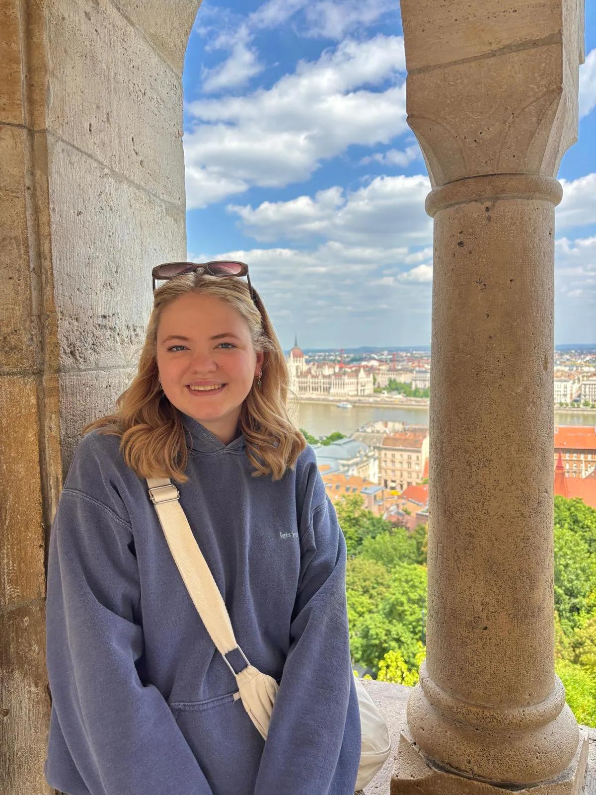 A young woman in a hoodie and sunglasses is stood in front of some stone columns and a view of a town and river.