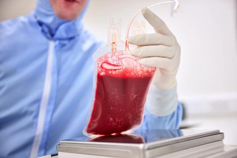 Scientist holding a cord blood unit in a laboratory environment above a scale. Wearing blue protective coverall suit.