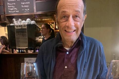 a man sat at a restaurant table smiling in front of a bowl of pasta. he is wearing a maroon shirt and blue jacket