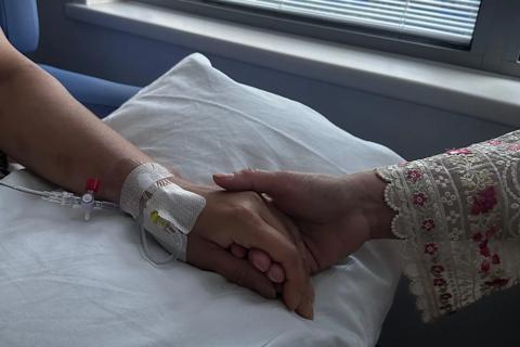 two hands holding on top of a white pillow, the hand on the left has a IV cannula below the wrist