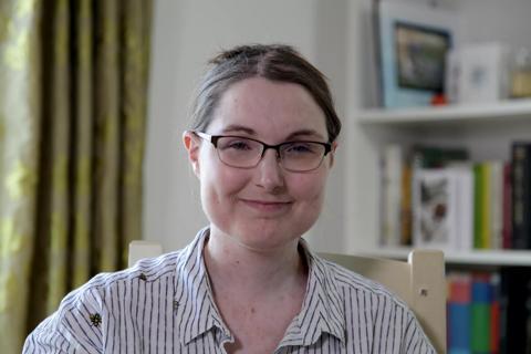 A white woman wearing glasses sits in her living room and smiles at the camera