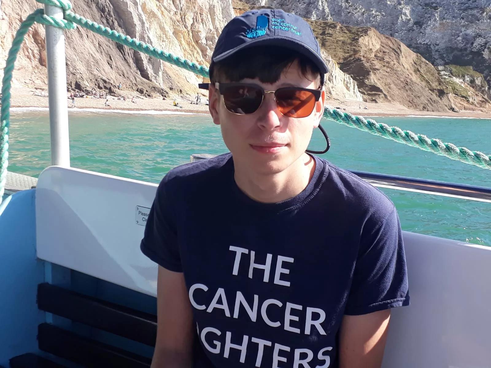 A young man wearing a cap and sunglasses. His t-shirt reads: the cancer fighters