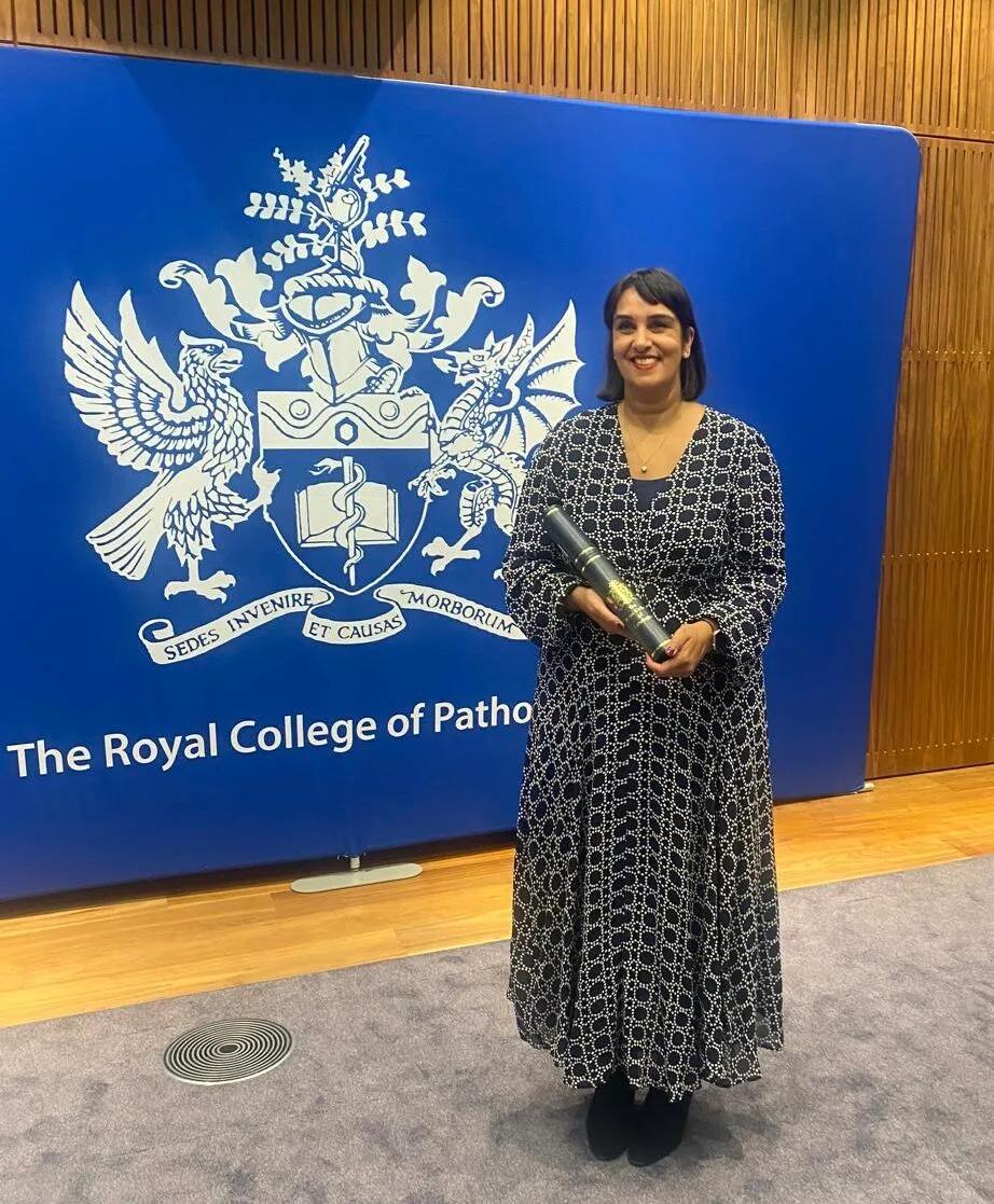 A smiling person holding a certificate in front of a Royal College of Pathologists display