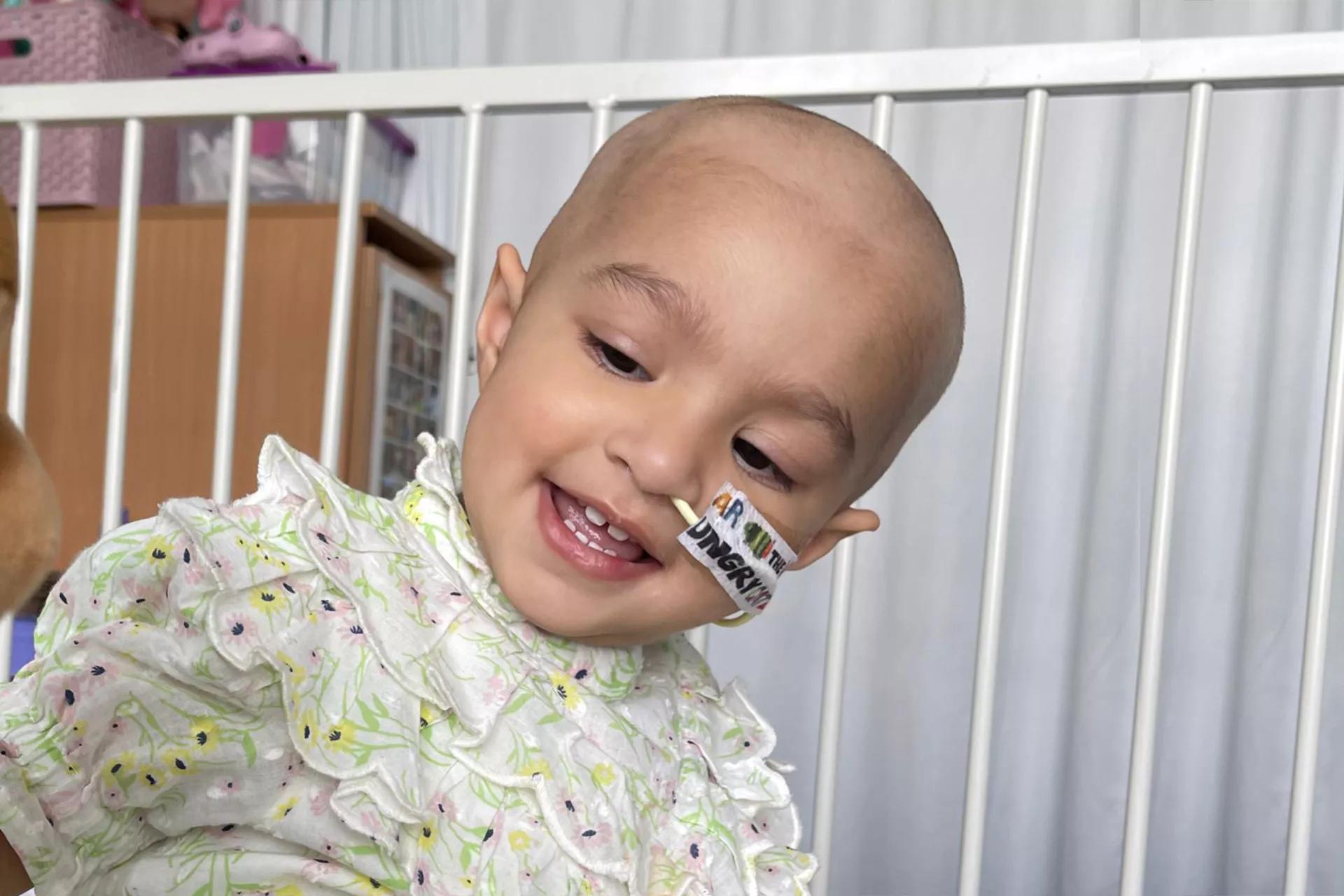 A toddler in a hospital cot smiling, she has an NG tube and no hair