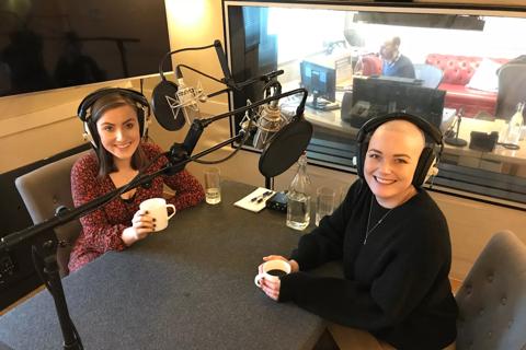 Two women sitting in a reording studio with mics hanging in front of them and earphones over each of their ears.