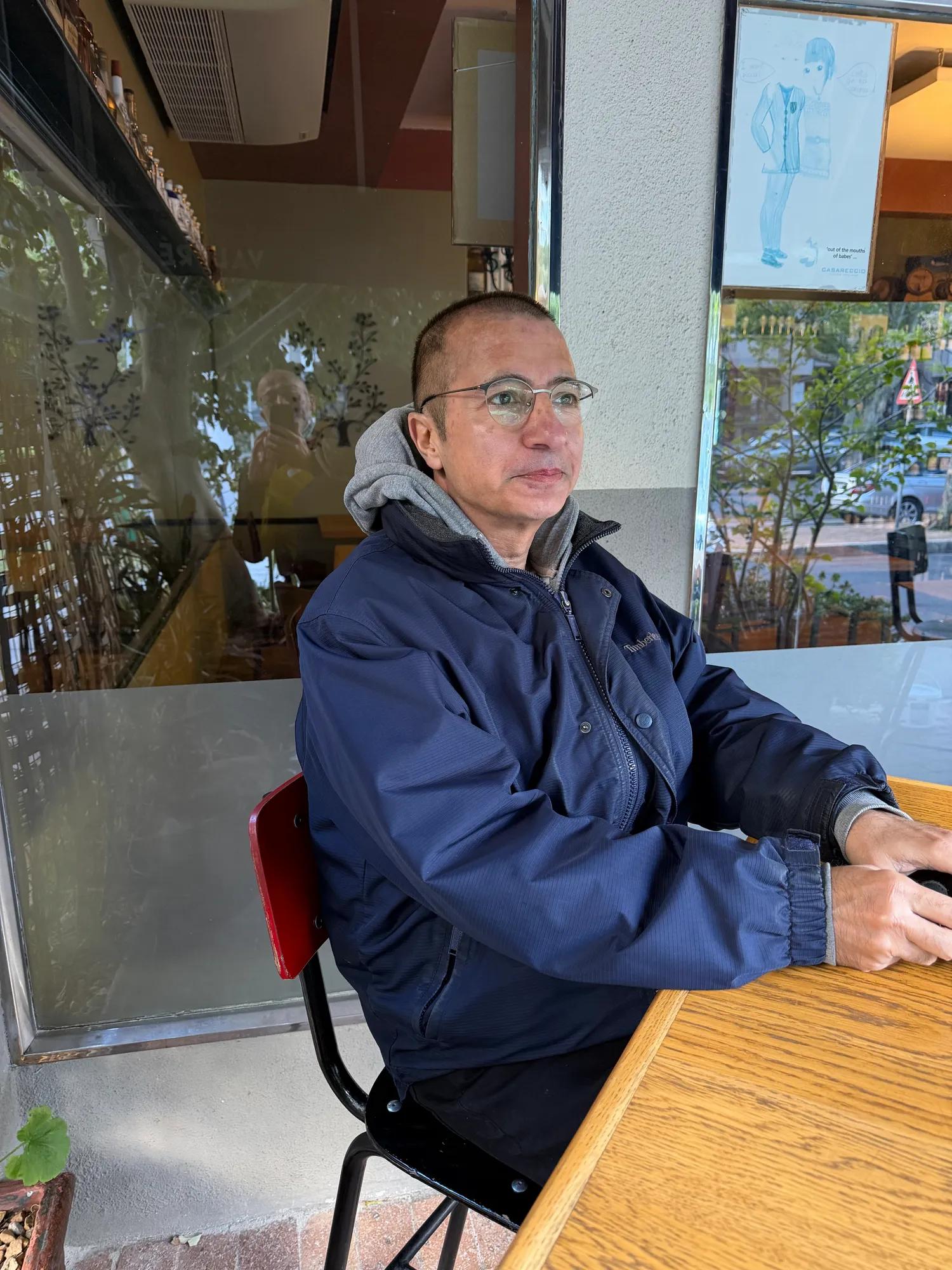 A man wearing glasses and a rain coat sits at a table outside a cafe