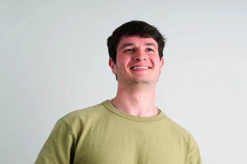 A 22 year old white male in a beige tshirt smiling, looking at camera