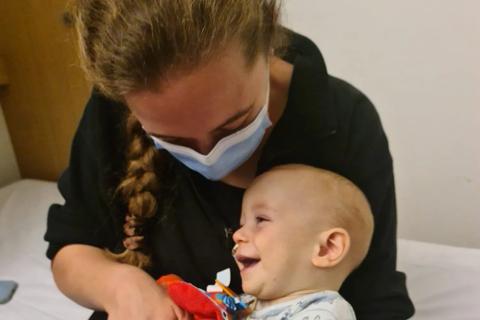 A baby boy sits on his mum's lap laughing. His mum tickles his tummy. He is holding a red babies toy and a blue sippy cup. His mum is wearing a hospital facemask.