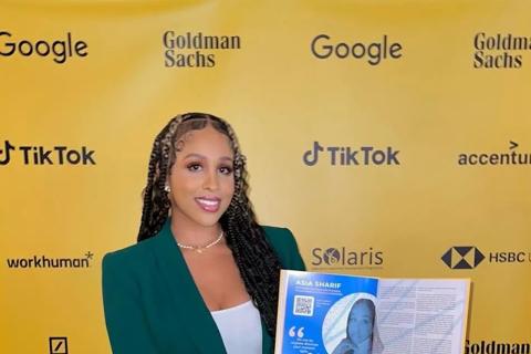 Asia, a young woman with long braided brown hair and green suit, holding open a booklet with her photo, in front of a yellow backgroun