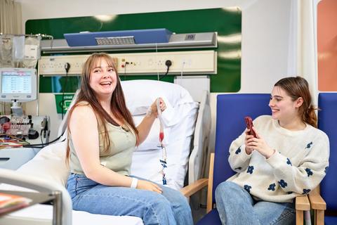 Donor holds stem cell bag sitting on the hospital bed,  while friend takes photo of her.
