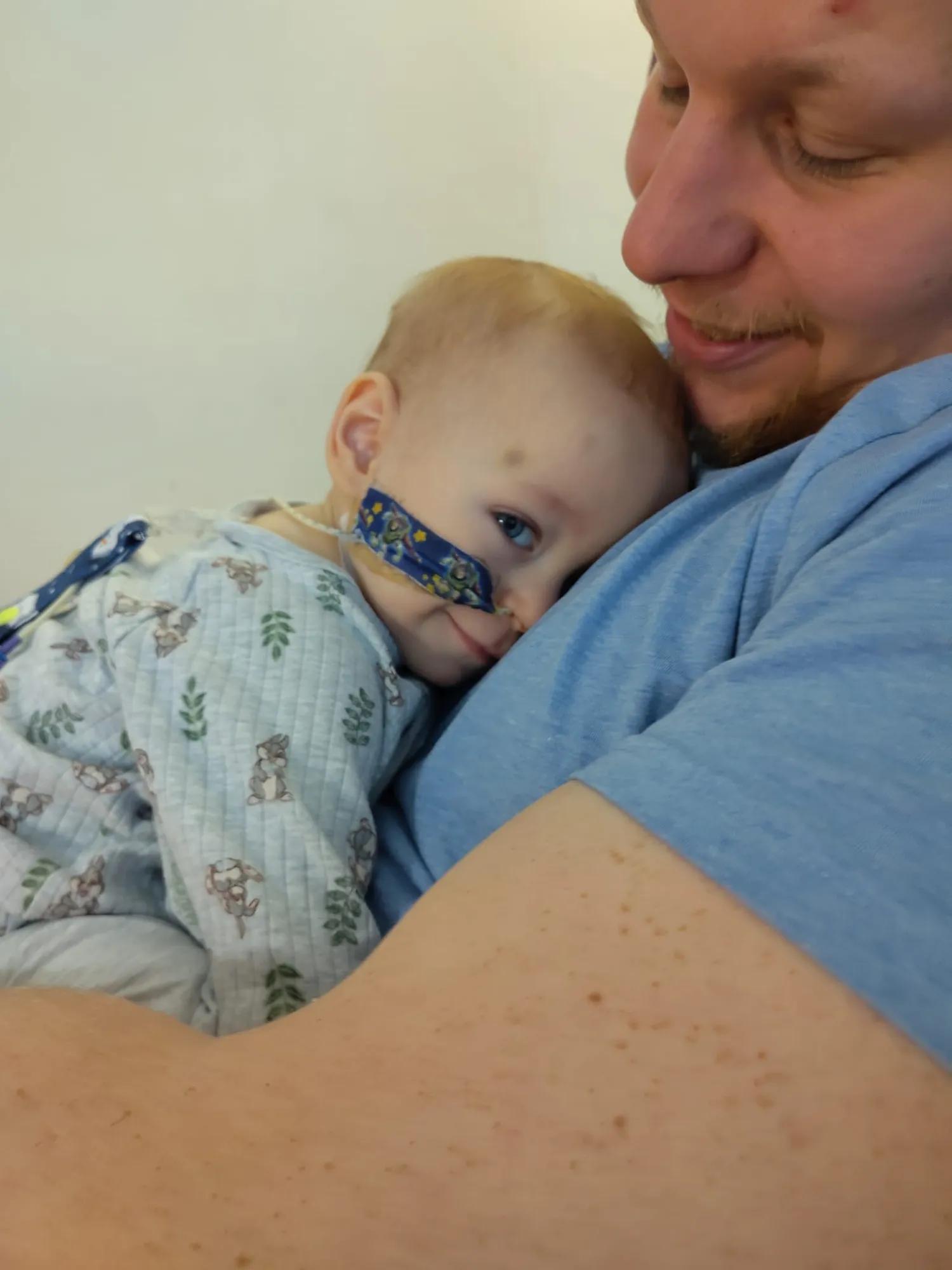 A baby boy is lying on his Dad's chest as his Dad smiles down at him. The baby boy has a feeding tube in his nose attached to his cheek with tape