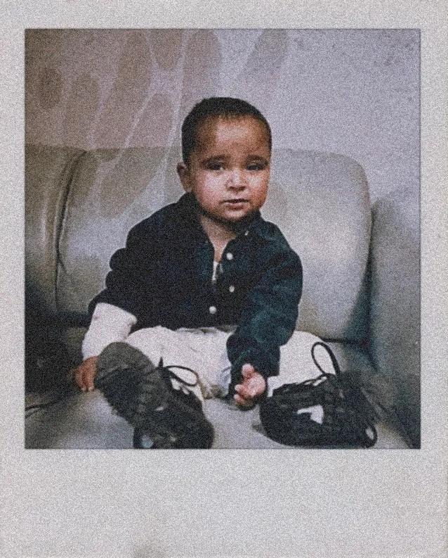 a polaroid photo of a baby boy on a grey sofa, he's wearing a black top and white trousers