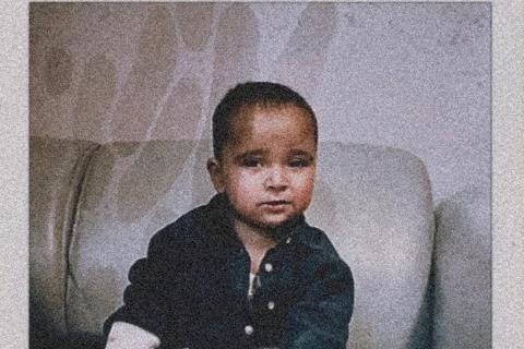 a polaroid photo of a baby boy on a grey sofa, he's wearing a black top and white trousers