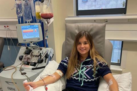 A young woman laying in a hospital bed attached to a machine taking blood, she is wearing a black t-shirt and trousers, she has long blonde hair and is smiling