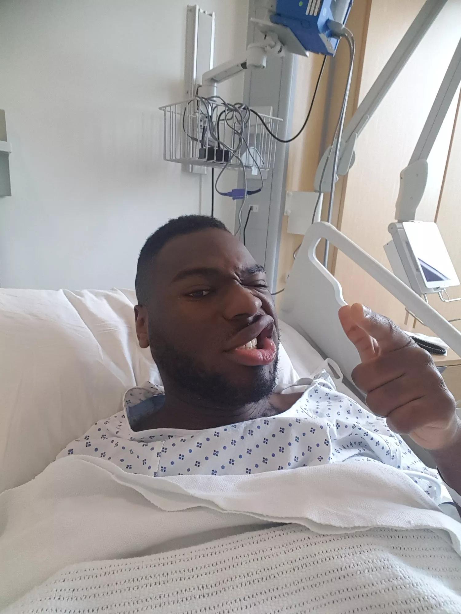 Carl in a hospital bed in his surgery gown pointing at the camera
