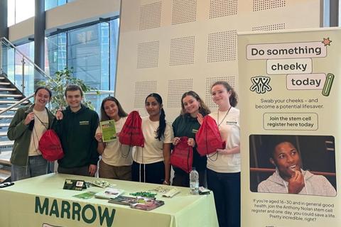 a group of young people are stood behind a donor recruitment stand, they are holding up leaflets and bags.