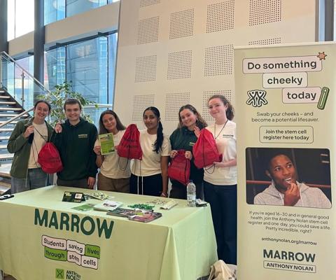 a group of young people are stood behind a donor recruitment stand, they are holding up leaflets and bags.