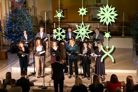 A choir stands on stage in front of a seated audience. Behind the choir is a Christmas tree