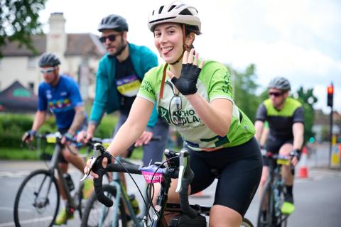 A woman wearing a helmet and waving while riding a bike with People cycling on bikes wearing their Anthony Nolan Ride London Shirt