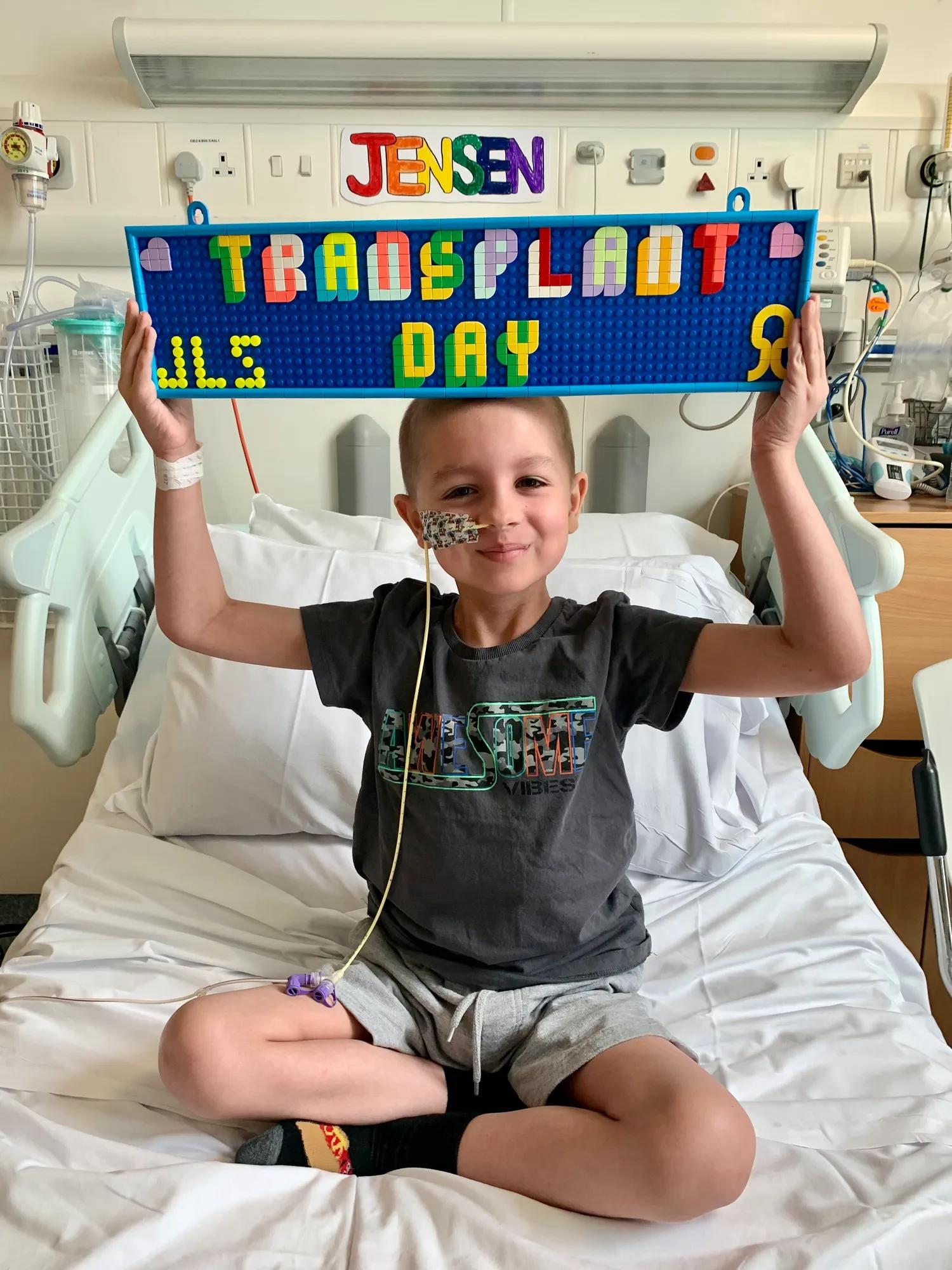 Eight year old, Jensen, sitting on a hospital bed proudly holding up a sign above his head that reads 'transplant day'