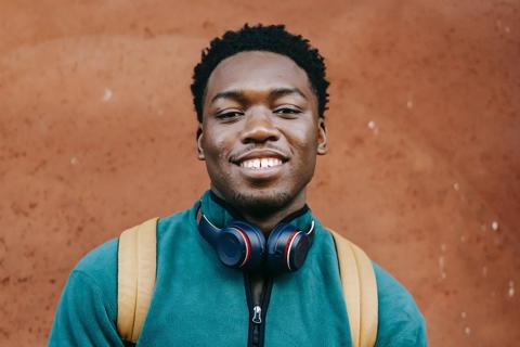 A young black man smiling. He is wearing a green jumper, a rucksack, and headphones around his neck.