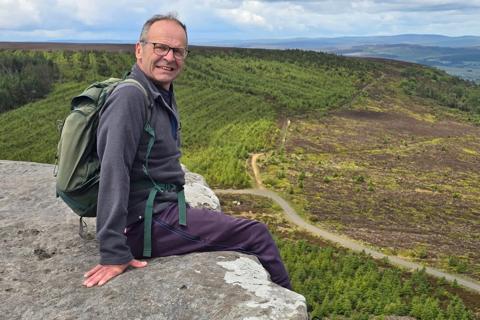 Richard sat on a rock at the top of a hill wearing a rucksack, smiling at the camera