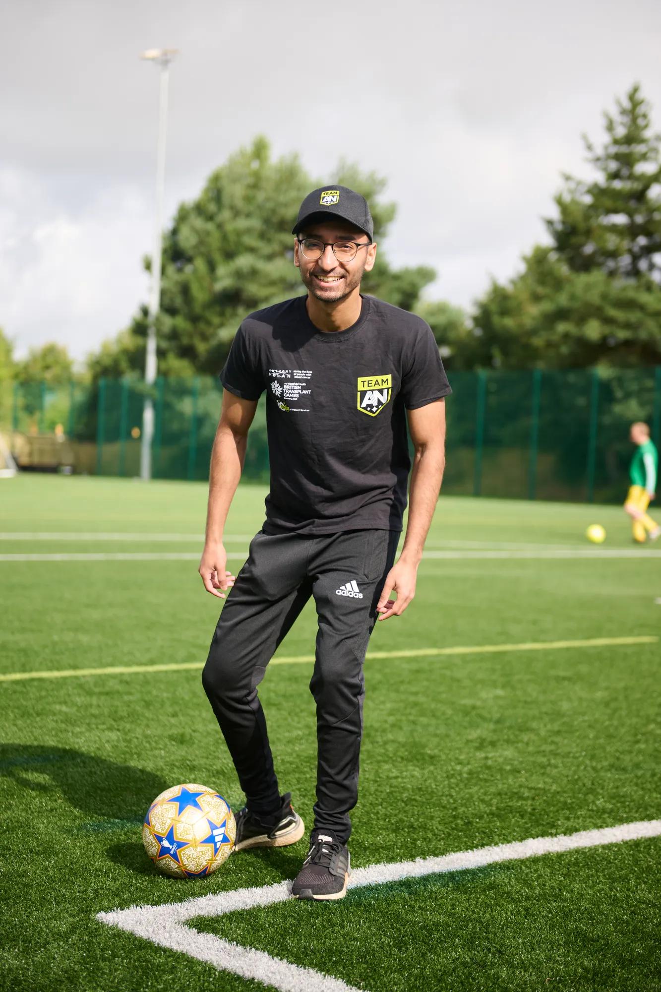 a young indian man wearing all black and an Anthony Nolan cap and t-shirt, kicking a football 