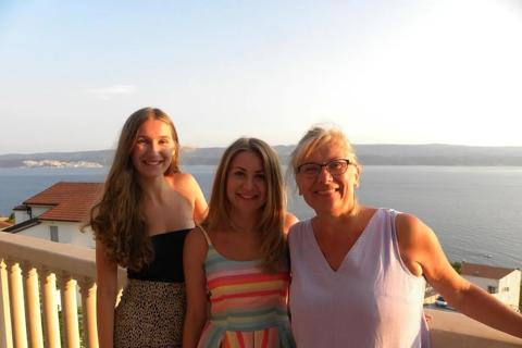 Kate smiling at the camera with her family, on a balcony on holiday with the sea in the background