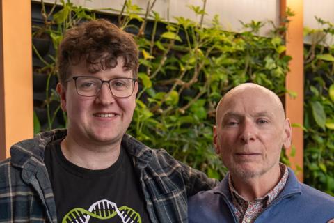 Two men, Tom (l) and Rod (r). Tom has his arm around, Rod. Tom is wearing an Anthony Nolan donor t-shirt under a shirt & blue jeans. Rod is wearing a floral shirt underneath a blue sweater and blue jeans.