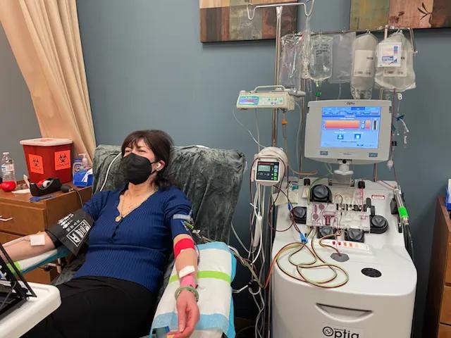 a woman in a hospital  hooked up to a machine through wires in her arm, she has brown hair and has a blue top, black trousers and a black face mask