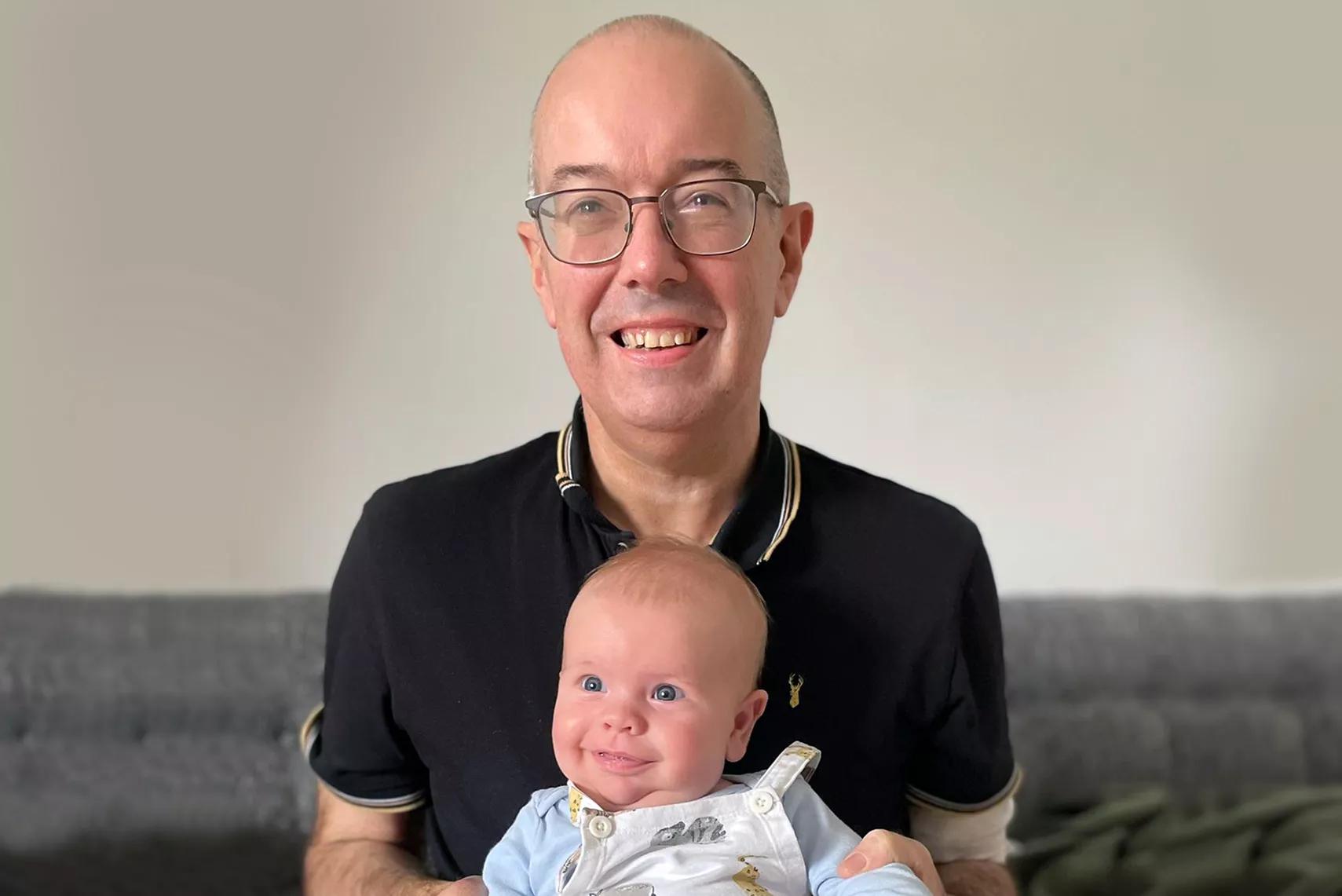 middle aged man with glasses, sitting on the sofa with a baby on his knee, he is smiling