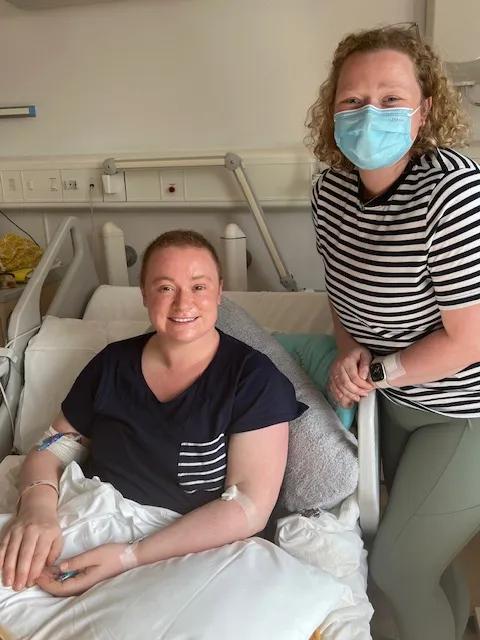 Emma, a young women with blonde curly hair, with her sister who is sitting in a hospital bed