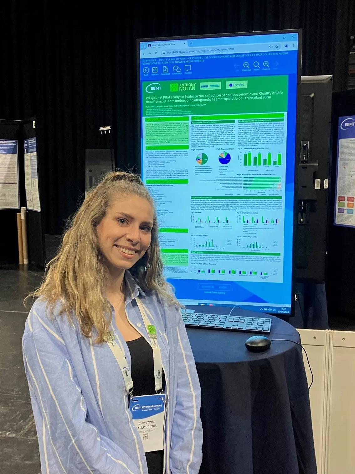 A whie woman wearing a denim overshirt with a smile on her face is standing in front of a poster