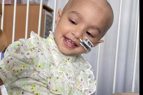 A toddler in a hospital cot smiling, she has an NG tube and no hair