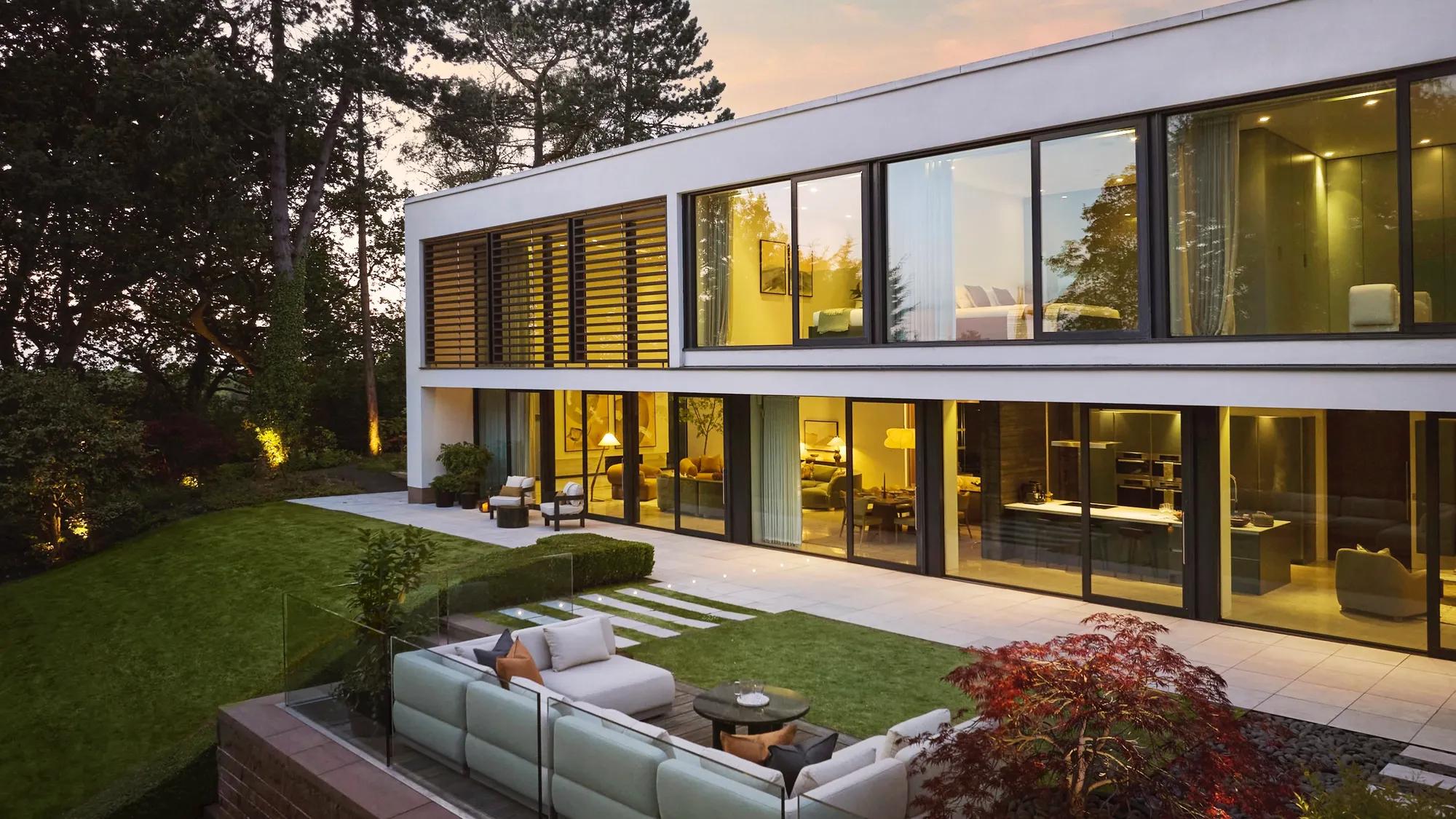 A photo of the house taken from the garden at sunset. The house is lit up inside with warm lighting and you can see into the rooms on both floors through the large windows. In the foreground is the garden furniture.
